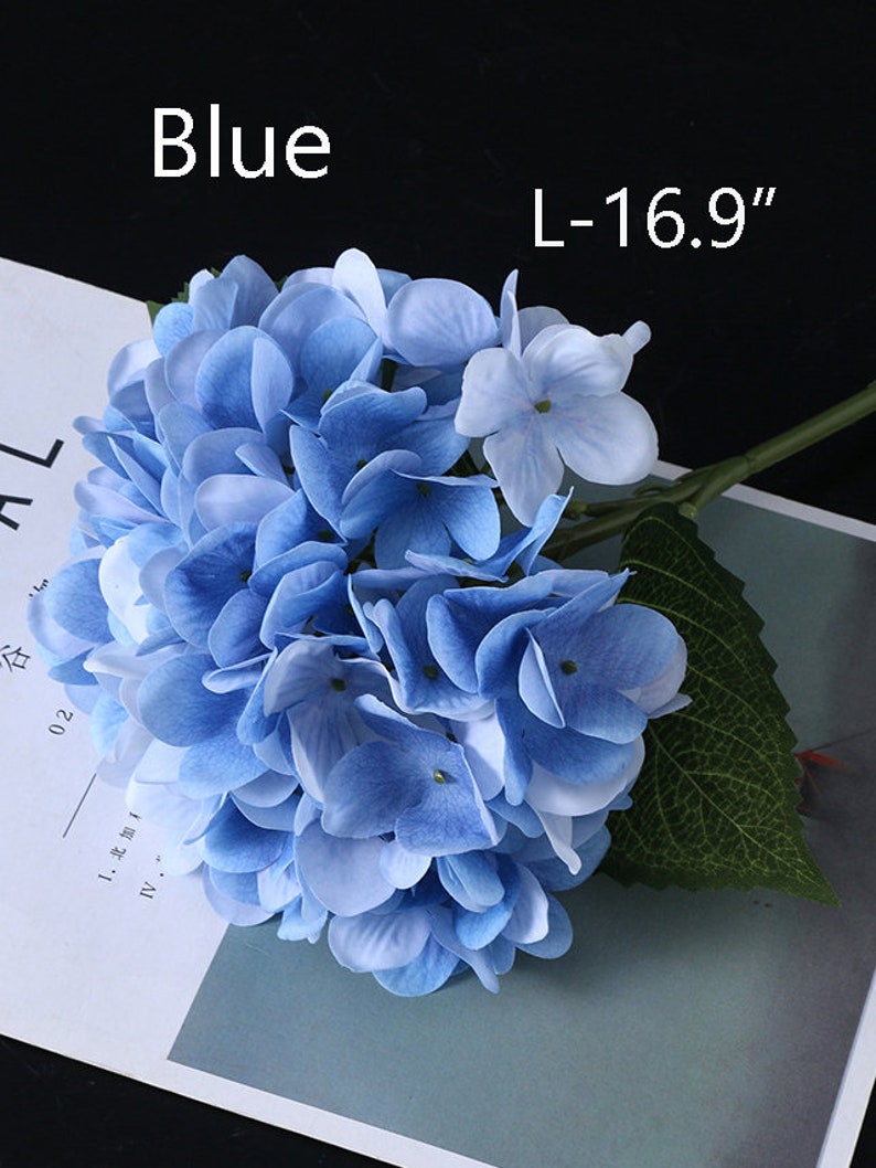Puede incluir: Una flor de hortensia azul con tallo y hoja verde. La flor est&aacute; etiquetada como "Azul" y mide L-43 cm. Los p&eacute;talos de la flor son una mezcla de azul claro y oscuro.