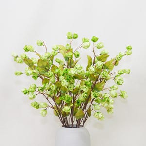 Puede incluir: Un ramo de flores artificiales con capullos y hojas verdes. Los tallos son marrones y las flores están en un jarrón blanco. El fondo es una pared blanca lisa.