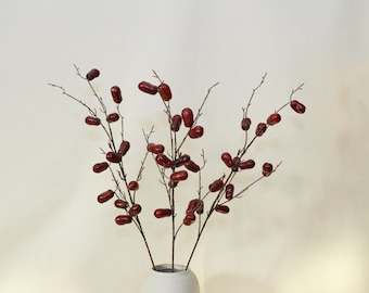 May include: A bouquet of artificial branches with red berries in a white vase. The branches are arranged in a vase on a white background.