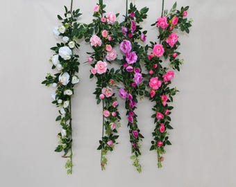 Puede incluir: Cuatro guirnaldas florales con rosas en tonos blanco, rosa y morado, acentuadas por hojas verdes. Las guirnaldas están suspendidas sobre un fondo neutro, creando una exhibición decorativa. Las rosas varían en tamaño e intensidad de color.