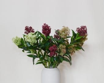 Puede incluir: Un ramo de flores artificiales en un jarrón blanco. Las flores tienen hojas verdes y racimos de pequeñas flores en tonos burdeos, crema y verde. El jarrón tiene una forma simple y redondeada.
