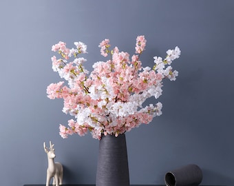 May include: A bouquet of pink and white artificial cherry blossoms in a gray vase. The vase is on a table with a white deer figurine.