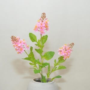 May include: A floral arrangement featuring pink flowers with yellow accents and green leaves. The flowers are in a white vase. The arrangement is set against a neutral background.