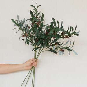 May include: A bouquet of artificial olive branches with green leaves and small, dark purple olives.