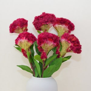 May include: A bouquet of red and green artificial flowers in a white vase. The flowers have a fuzzy texture and are arranged in a vase with a textured surface.
