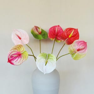 May include: A collection of seven artificial anthurium flowers in a light gray vase. The heart-shaped spathes feature a variety of colors including solid white, pale pink, vibrant red, and variegated combinations of green, pink, and red.