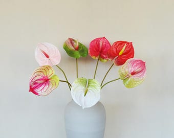 Puede incluir: Un ramo de siete flores de anturio artificiales en un jarrón gris claro. Las espatas en forma de corazón presentan una variedad de colores que incluyen blanco sólido, rosa pálido, rojo vibrante y combinaciones de verde, rosa y rojo.