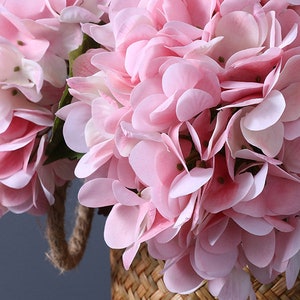 Puede incluir: Primer plano de un ramo de flores de hortensia rosa en una cesta tejida. Las flores tienen m&uacute;ltiples capas de delicados p&eacute;talos en varios tonos de rosa. La cesta est&aacute; hecha de material tejido natural y se ve un asa de cuerda.