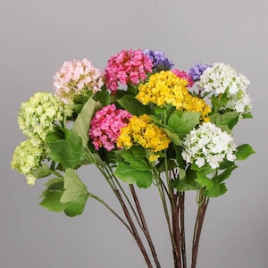 May include: A bouquet of artificial flowers with various colors, including white, yellow, pink, and purple. The flowers are arranged in a cluster and have green leaves.