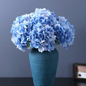 Puede incluir: Un ramo de flores de hortensia azul en un jarr&oacute;n turquesa texturizado. Las flores tienen m&uacute;ltiples capas de p&eacute;talos en tonos azul claro y medio. El jarr&oacute;n es de color s&oacute;lido con una apariencia agrietada.