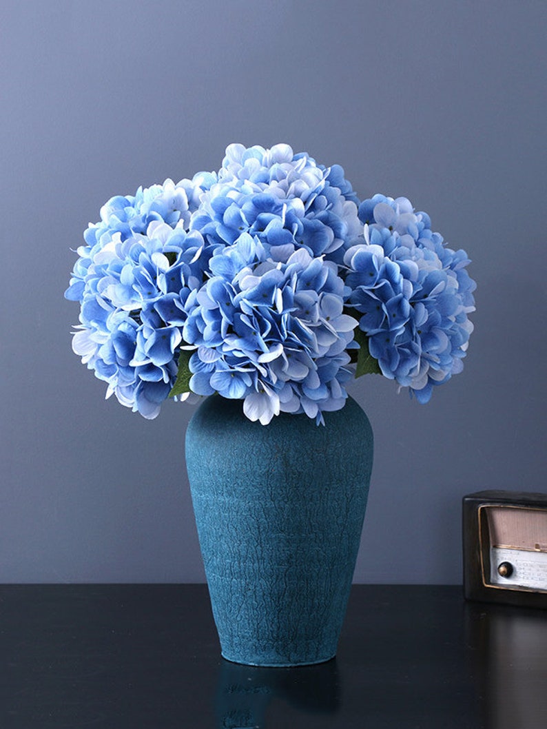 Puede incluir: Un ramo de flores de hortensia azul en un jarr&oacute;n turquesa texturizado. Las flores tienen m&uacute;ltiples capas de p&eacute;talos en tonos azul claro y medio. El jarr&oacute;n es de color s&oacute;lido con una apariencia agrietada.