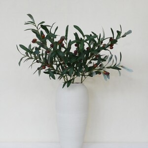 May include: A white ceramic vase with a textured surface holds a bouquet of green olive branches with small brown olives.