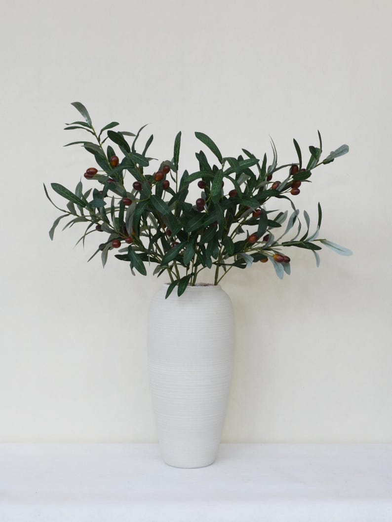May include: A white ceramic vase with a textured surface holds a bouquet of green olive branches with small brown olives.