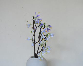 May include: A floral arrangement featuring delicate, light purple flowers with yellow centers and green accents. The flowers are arranged on dark brown stems, displayed in a white, textured vase. The background is a plain, off-white wall.