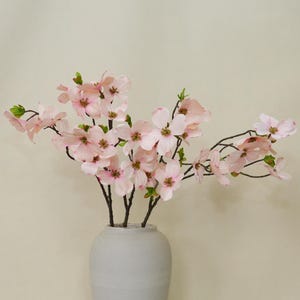 Puede incluir: Un ramo de flores artificiales de cornejo en un jarrón de cerámica blanco. Las flores son de color rosa pálido con centros amarillos y detalles verdes. El jarrón tiene una superficie texturizada y una forma redonda. El fondo es de color neutro.