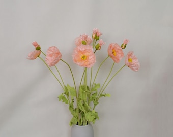 May include: A bouquet of peach-colored silk poppy flowers with green stems and leaves in a gray ceramic vase.