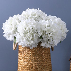 Puede incluir: Una cesta tejida llena de flores de hortensia blancas. La cesta est&aacute; hecha de material tejido de color natural con asas de cuerda. Las flores est&aacute;n agrupadas, creando una apariencia completa y texturizada sobre un fondo azul.