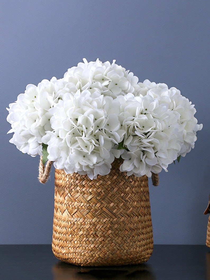 Puede incluir: Una cesta tejida llena de flores de hortensia blancas. La cesta est&aacute; hecha de material tejido de color natural con asas de cuerda. Las flores est&aacute;n agrupadas, creando una apariencia completa y texturizada sobre un fondo azul.