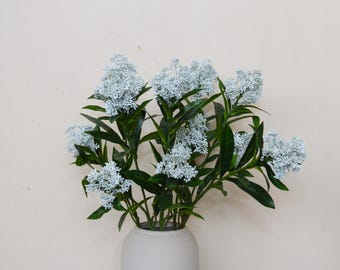 Puede incluir: Un ramo de flores azul claro con hojas verdes en un jarrón de cerámica blanco. Las flores tienen un aspecto texturizado y agrupado, y el jarrón tiene una forma simple y redondeada. El arreglo está sobre un fondo neutro.
