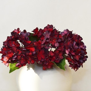 以下が含まれることがあります： 白のセラミック花瓶に入った、濃い赤と紫のアジサイの花束。 花は層状の外観で、緑の葉が見えます。 花瓶はテクスチャ加工された表面を持っています。