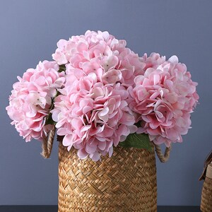Puede incluir: Una cesta tejida llena de flores de hortensia rosas. Las flores tienen m&uacute;ltiples capas de delicados p&eacute;talos. La cesta est&aacute; hecha de un material tejido marr&oacute;n claro con asas de cuerda. El fondo es de un gris azulado s&oacute;lido.