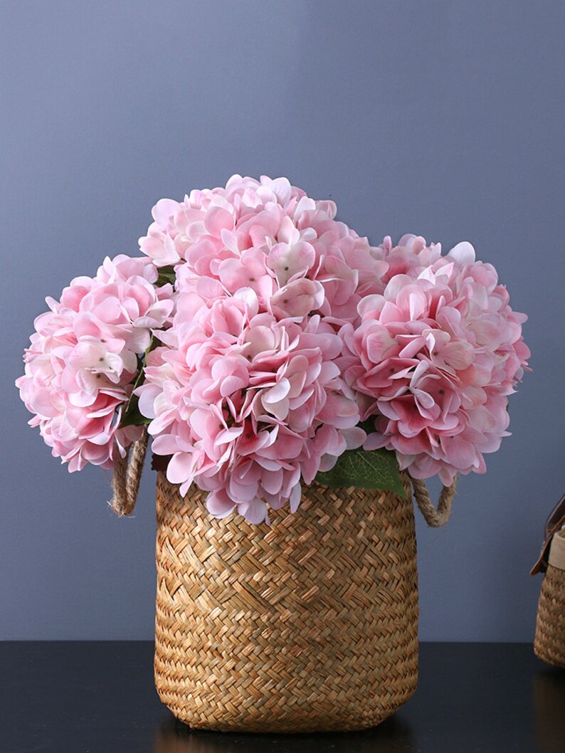 Puede incluir: Una cesta tejida llena de flores de hortensia rosas. Las flores tienen m&uacute;ltiples capas de delicados p&eacute;talos. La cesta est&aacute; hecha de un material tejido marr&oacute;n claro con asas de cuerda. El fondo es de un gris azulado s&oacute;lido.