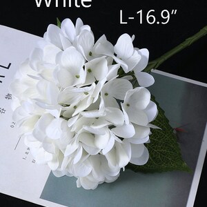 Puede incluir: Una flor de hortensia blanca con tallo y hoja verdes. El cabezal de la flor es un grupo de peque&ntilde;os y delicados p&eacute;talos. El texto "White" y "L-16.9" son visibles. La flor es un art&iacute;culo decorativo.