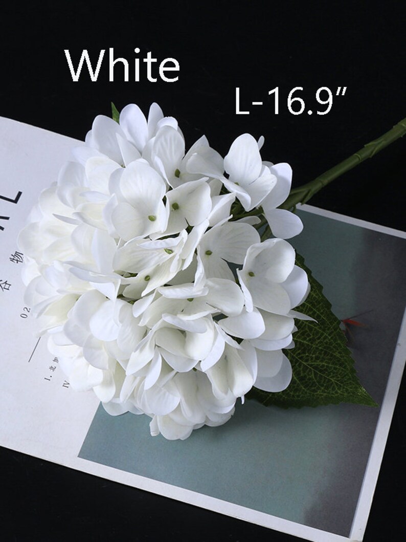 Puede incluir: Una flor de hortensia blanca con tallo y hoja verdes. El cabezal de la flor es un grupo de peque&ntilde;os y delicados p&eacute;talos. El texto "White" y "L-16.9" son visibles. La flor es un art&iacute;culo decorativo.