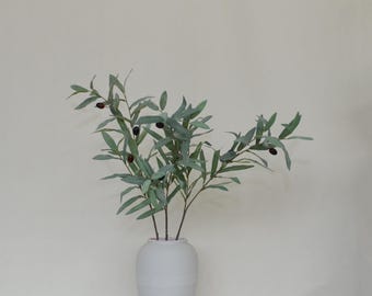 May include: A white ceramic vase holds several olive branches with green leaves and dark olives. The branches are arranged to create a natural, organic aesthetic. The vase is simple and cylindrical.