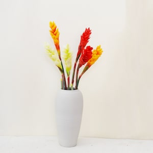 May include: A white ceramic vase with a textured surface holds a bouquet of artificial flowers. The flowers are a mix of yellow, orange, and red, and they are arranged in a symmetrical pattern.