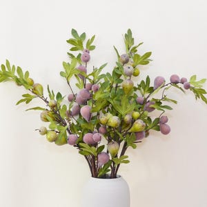 May include: A bouquet of artificial fig branches with green leaves and purple figs in a white ceramic vase.