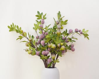 May include: A bouquet of artificial fig branches with green leaves and purple figs in a white ceramic vase.