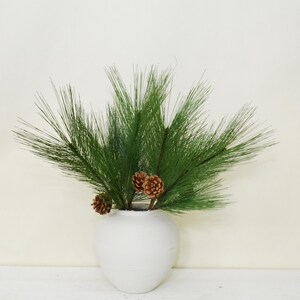 May include: A white ceramic vase holds a display of green pine branches and several brown pine cones. The arrangement is set against a neutral background, creating a simple, natural aesthetic. This decorative piece is suitable for home decor.