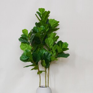 May include: A vibrant green artificial fiddle leaf fig plant. The plant features large, glossy, green leaves and slender brown stems. It is displayed in a white, rounded vase. The plant's height is approximately 4 feet.