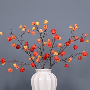 Op de afbeelding: Een witte keramische vaas met een textuur bevat een boeket van oranje en gele papieren bloemen. De bloemen zijn in een losse, natuurlijke stijl gerangschikt.