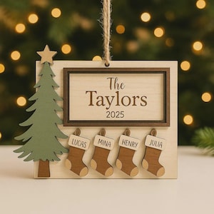 Puede incluir: Adorno navideño de madera con fondo de veta de madera clara. El adorno presenta un árbol de Navidad, el texto "The Taylors 2025" y cuatro calcetines con nombres. El adorno está suspendido por un cordel natural.
