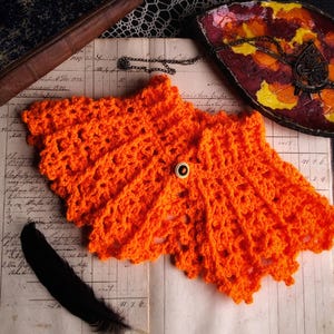 Könnte beinhalten: Leuchtend orange gehäkelter Schal mit Knopfverschluss. Der Schal hat ein strukturiertes Design und einen gewellten Rand. Er wird auf gealtertem Papier zusammen mit einer schwarzen Feder und einem antiken Buch präsentiert.