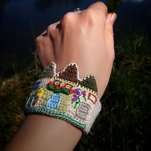 Könnte beinhalten: Ein weißes gehäkeltes Armband mit bunten Stickereien, die eine kleine Stadt mit Häusern, Blumen und Bäumen darstellen.