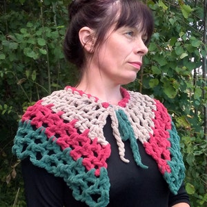 May include: A crocheted collar in shades of beige, pink, and green. The collar is made of a soft, fuzzy yarn and has a loose, open weave. It is worn by a person with dark hair and a black shirt.