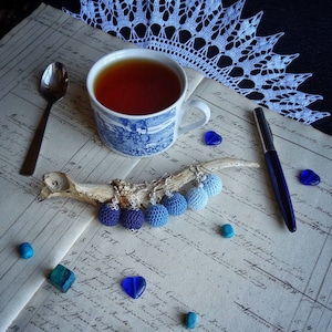 May include: A set of blue crocheted earrings with silver ear wires, displayed on a bone-like object. The earrings are on a vintage-looking ledger with a blue pen, a cup of tea, and blue glass hearts.