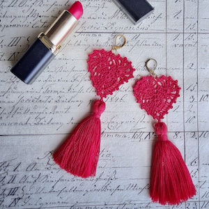 May include: Two pink crocheted heart earrings with tassels. The earrings are on a white surface with handwritten text.