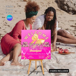 May include: A pink sign on a wooden easel with the words "WELCOME Women's PRAYER Breakfast" in white and gold. The sign is decorated with floral accents and a yellow bow. Two women are seated on a blanket in the background.