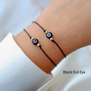 Op de afbeelding: Twee zwarte armbandjes van touw met een zwarte boze oog bedel op elk. Het armbandje aan de linkerkant heeft een zilveren kraal en het armbandje aan de rechterkant heeft een gouden kraal. De tekst "Black Evil Eye" is zichtbaar onder de armbandjes.