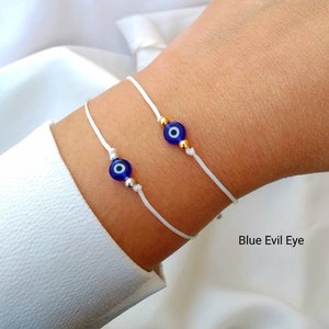 May include: Two white string bracelets with blue evil eye charms. The charms have a gold rim. The bracelets are on a person's wrist. The text "Blue Evil Eye" is visible below the bracelets.