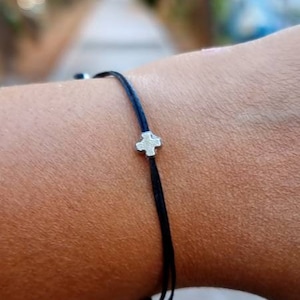 Könnte beinhalten: Ein schwarzes Armband aus Schnur mit einem silbernen Kreuzanhänger.