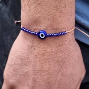 May include: A blue evil eye charm bracelet with a brown cord. The bracelet is made of small blue beads and features a blue evil eye charm with a white centre.