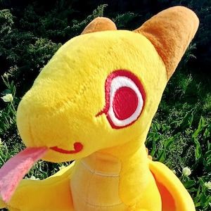 May include: A yellow plush toy shaped like a dragon with large red eyes, a light pink forked tongue, and brown accents on the head. The creature features small wings, a textured fabric body, and a pointed tail, presented in a soft, stuffed design.