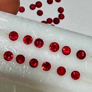 Pode incluir: Uma coleção de pequenas gemas redondas, de cor vermelha vibrante. As gemas estão espalhadas sobre uma superfície branca, algumas dispostas em linha. As gemas têm uma superfície polida e refletora, sugerindo que são cortadas e facetadas. A imagem está bem iluminada.