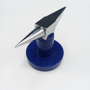 Puede incluir: Un pequeño yunque de metal plateado con un cuerno puntiagudo y una parte superior plana. El yunque está montado sobre una base redonda de color azul oscuro. El yunque es una herramienta utilizada para dar forma al metal.