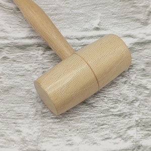Könnte beinhalten: Ein Holzhammer mit hellbrauner Oberfläche und einem abgerundeten Kopf. Der Hammer ist aus Naturholz gefertigt und hat eine glatte Oberfläche.